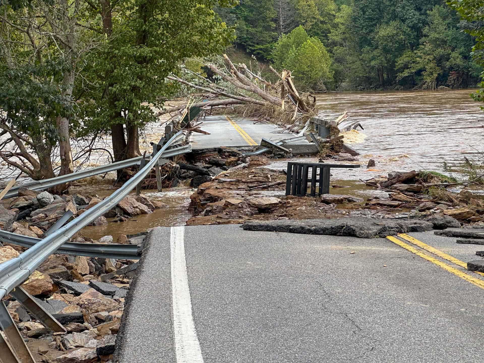Flooded road with fallen trees and damaged guardrail due to flood risk.